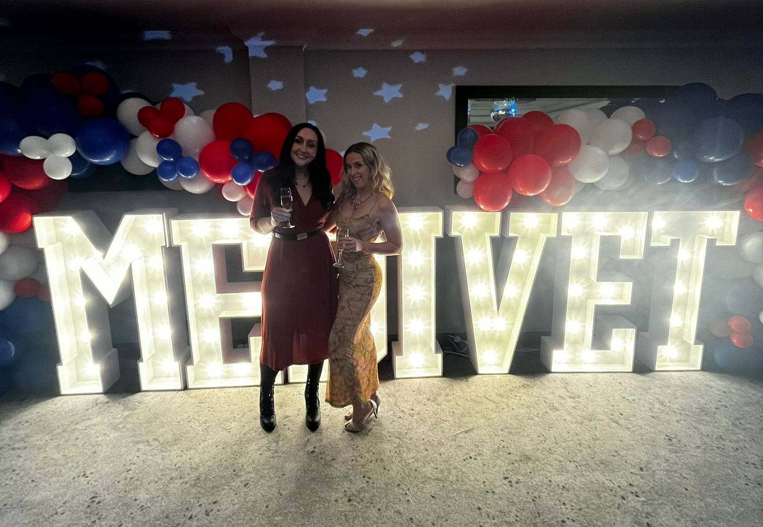 Two women smiling with drinks stand before lit "MEIVET" letters. Red, white, and blue balloons decorate the background. Joyful mood.