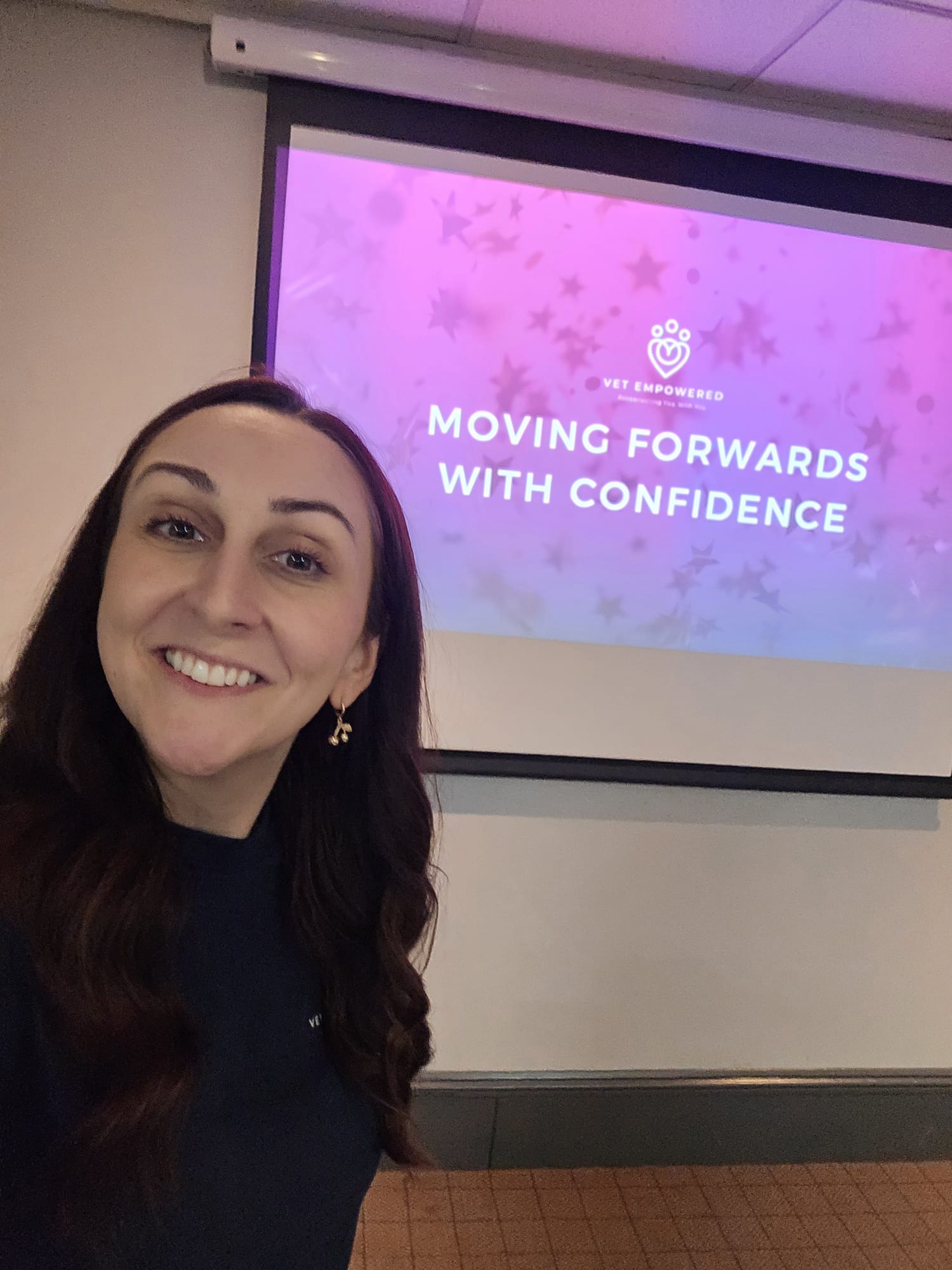 Smiling woman in front of a screen showing "Moving Forwards with Confidence" on a purple background with stars in a conference room.