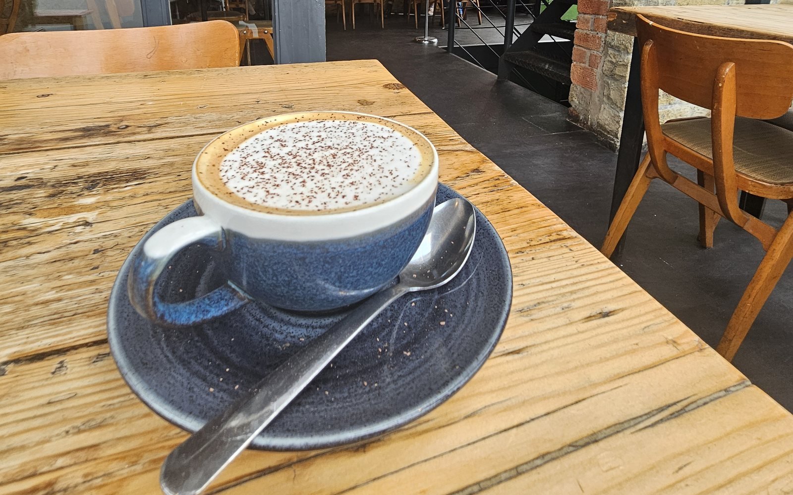 Cappuccino in a blue cup on a wooden café table, with empty chairs behind
