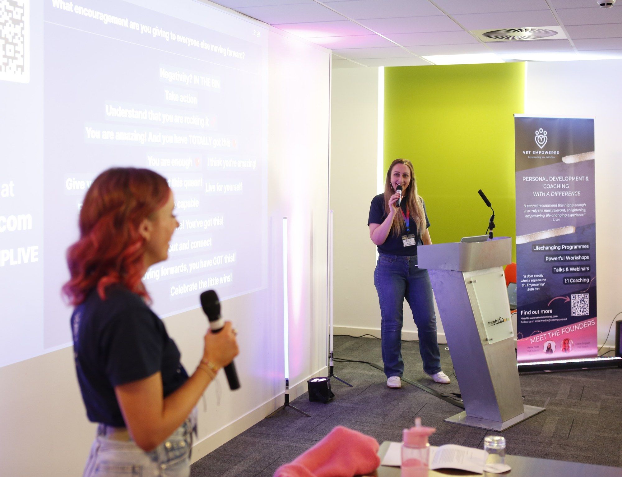 Two women with microphones present in a conference room. A screen displays motivational text. A banner promotes a coaching program.