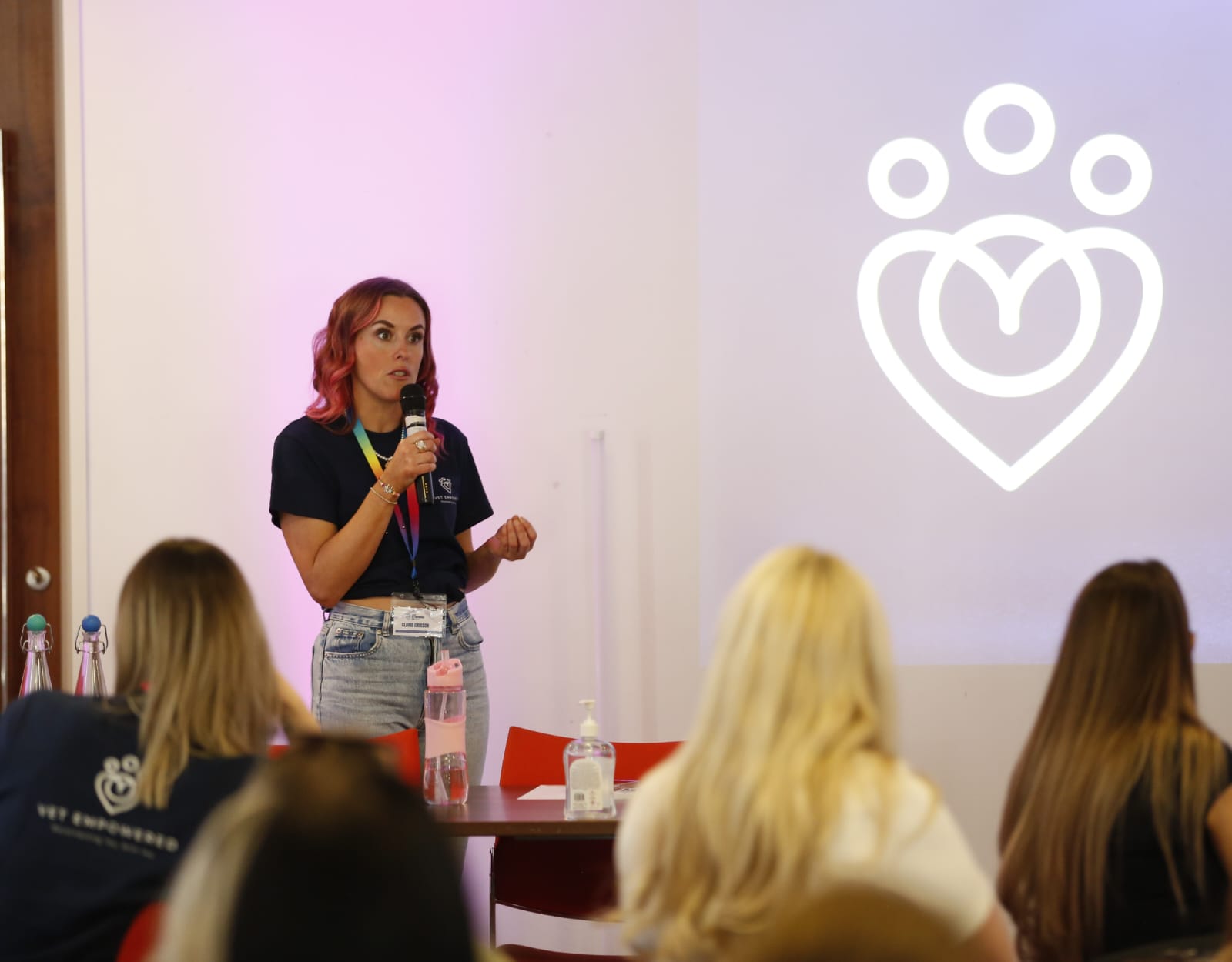A woman speaks to an audience, holding a microphone. A heart logo is projected behind her. The room has red chairs and a purple light.
