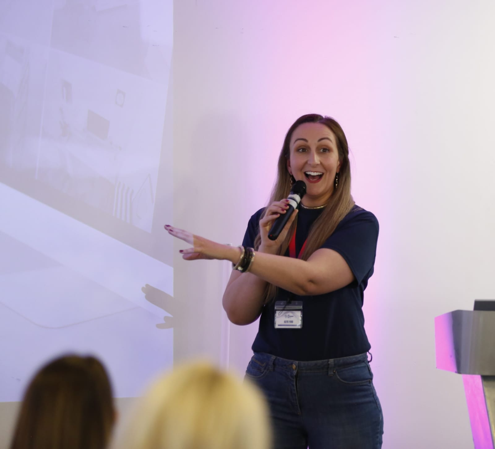 Woman speaking enthusiastically into a microphone, pointing at a projected screen. Casual attire, bright background, engaged audience.